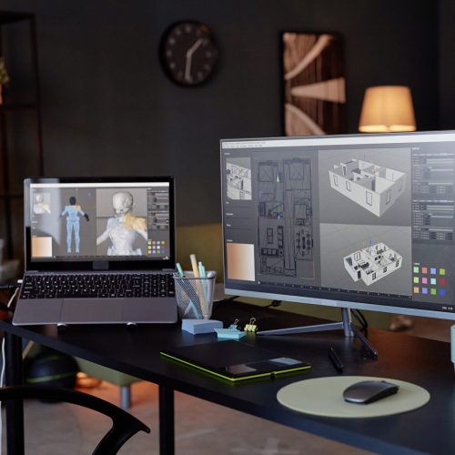 Modern office workspace with dual screens displaying 3D digital models and illustrations. Convenient office setup including stationery, coffee mug, and tech gadgets for efficient workflow